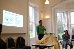 A tutor in a green shirt presenting a slide on Psychodynamic Counselling to an attentive audience at a Diploma Open Day, in an elegant room with tall sash windows.