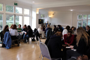 Prospective students seated in small groups, engaged in discussion activities during a Diploma Open Day in a bright, airy hall with large windows.