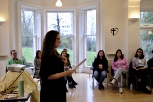 A presenter in black holding notes and addressing a seated audience during a session at a Diploma Open Day, in a room with a large bay window overlooking a garden.