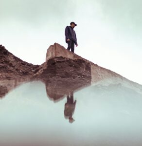 A person stands on a rocky ledge looking down at their reflection in still water below.