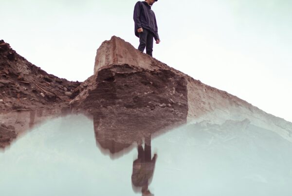 A person stands on a rocky ledge looking down at their reflection in still water below.