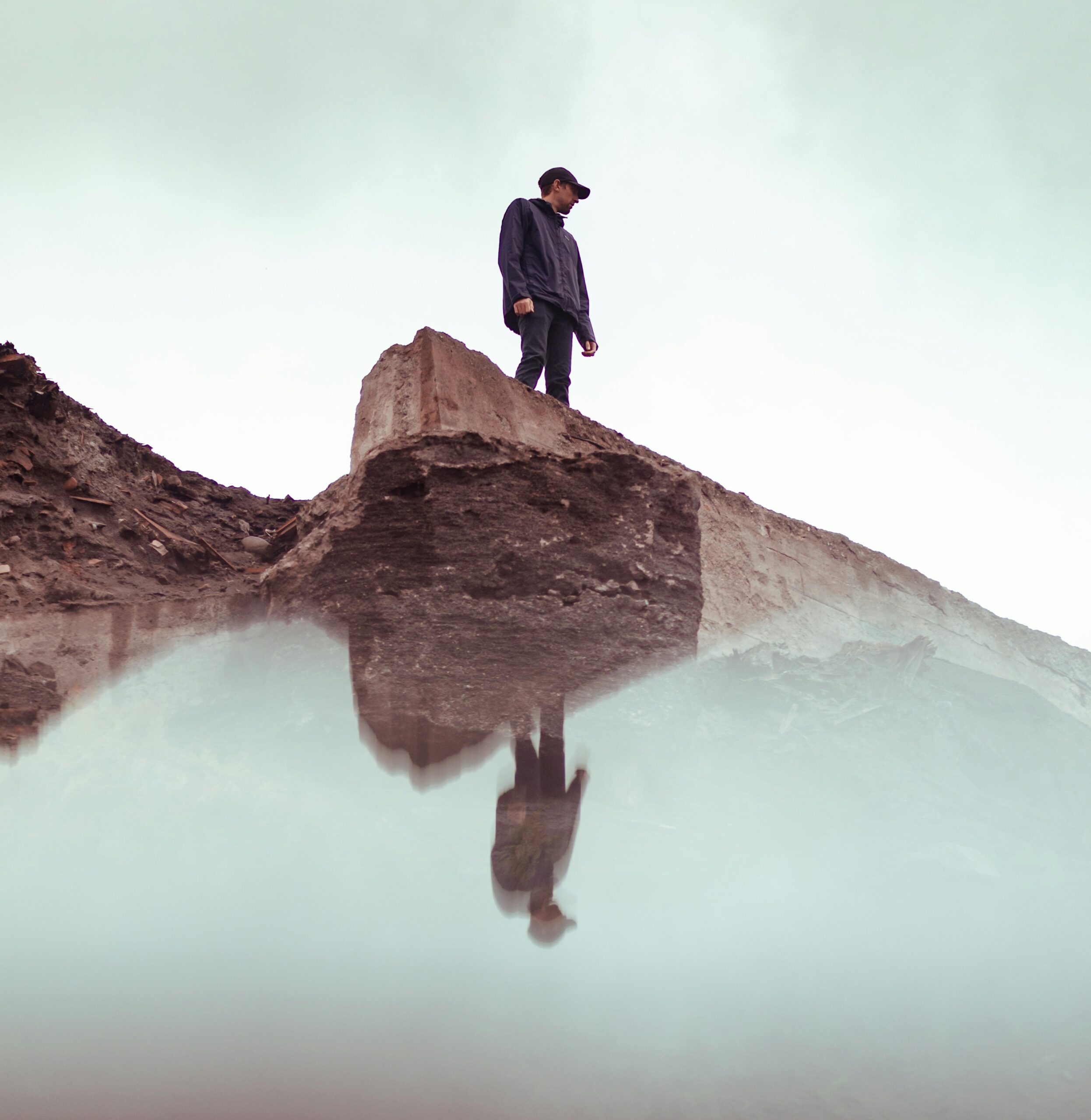 A person stands on a rocky ledge looking down at their reflection in still water below.