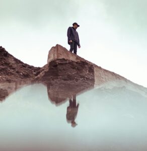 A person stands on a rocky ledge looking down at their reflection in still water below.