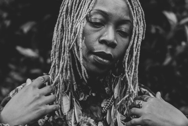 A black and white portrait of a woman with locs, eyes closed, set against a backdrop of leaves.